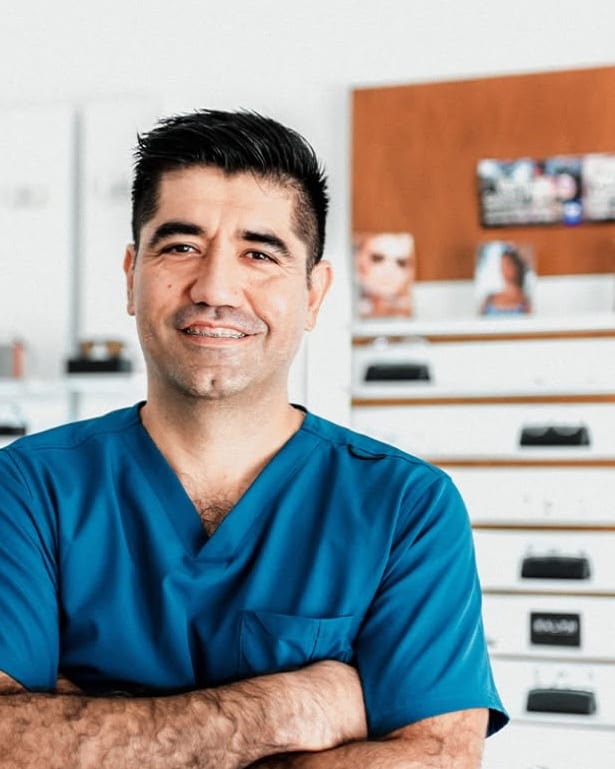 El Dr. Juan Antonio Liera García, sonriendo, con uniforme médico azul y el aparador de armazones de su óptica detrás